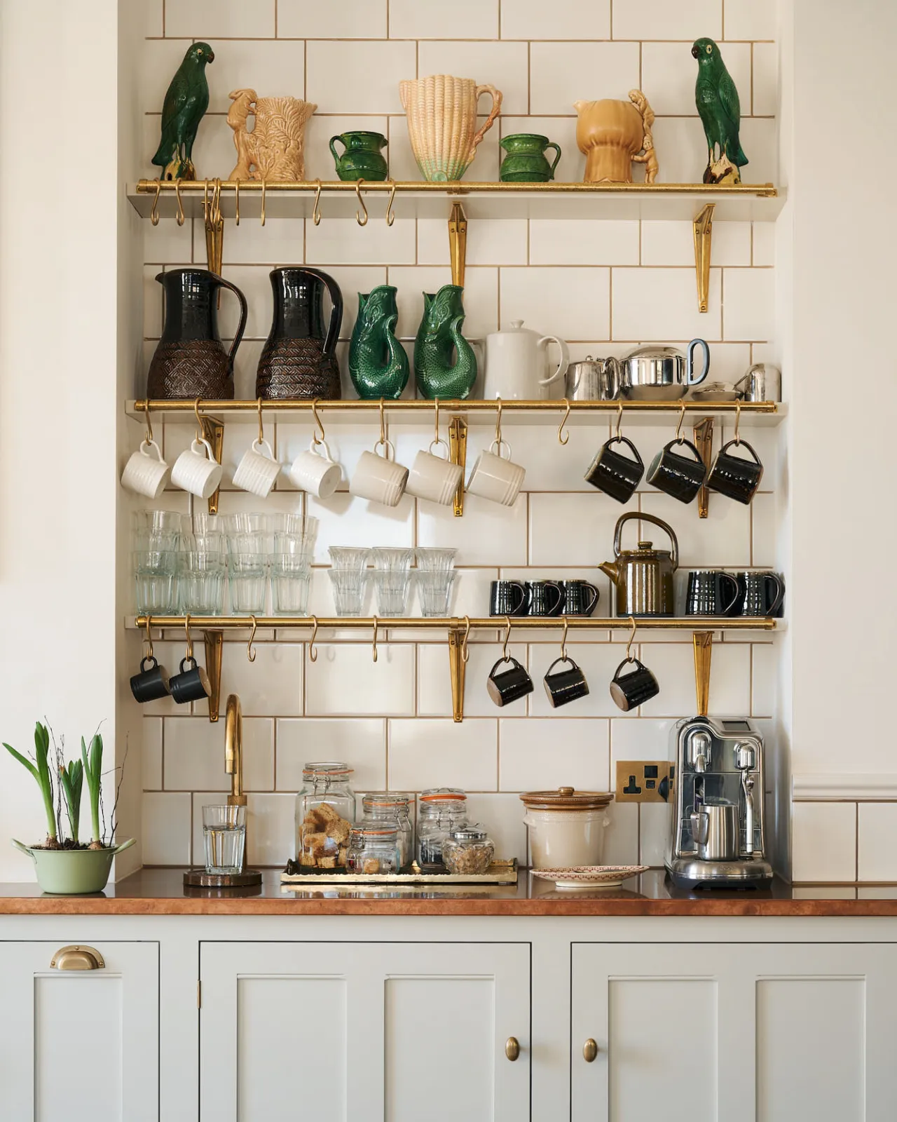 A little coffee-making nook with three fixed wall shelves, fitted with brass rails and adorned with ‘s’ hooks holding rows of glossy mugs. The entire image shines with reflections from the aged brass, glassware and ceramics.