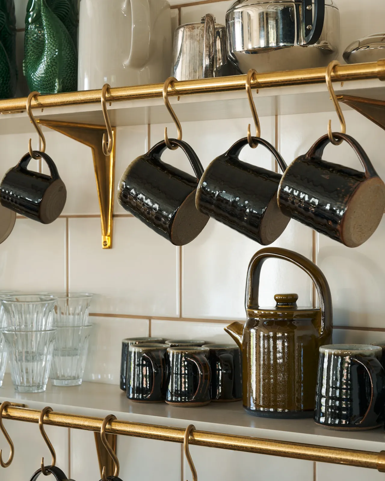Glossy handmade ceramic mugs hang delicately from ‘S’ hooks that are placed over an aged brass rail, fitted to a shelf at the back of a little drinks-making station. Aged brass and amber-coloured ceramics contrast with a cream-tiled wall, creating a warm, glowy feel.
