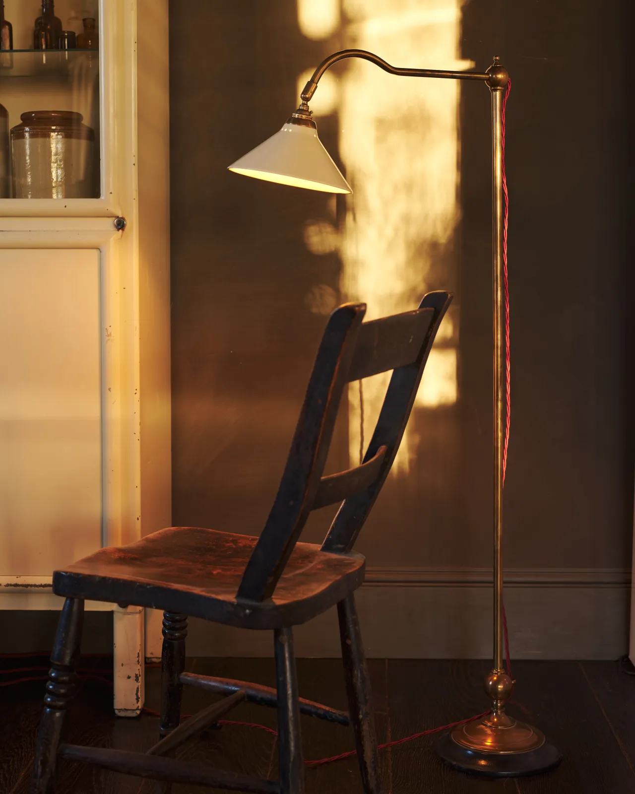 a front on shot of the grand tour reading lamp by a wooden chair, shot during sunset golden hour, lighting the wall with its warm light.