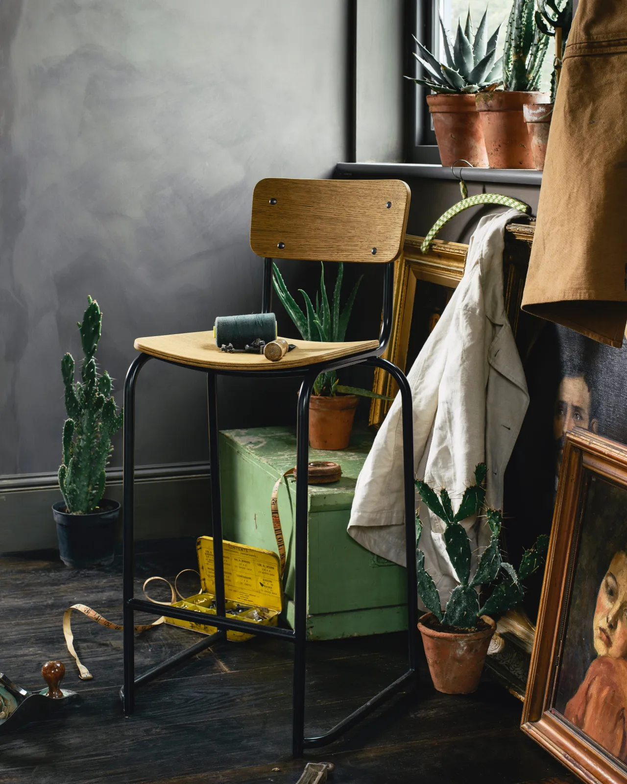 An oblique view of a mid-century-style industrial bar stool with a black tubular metal frame and shaped oak veneer plywood seat and backrest in a dark room with antique paintings and furniture.