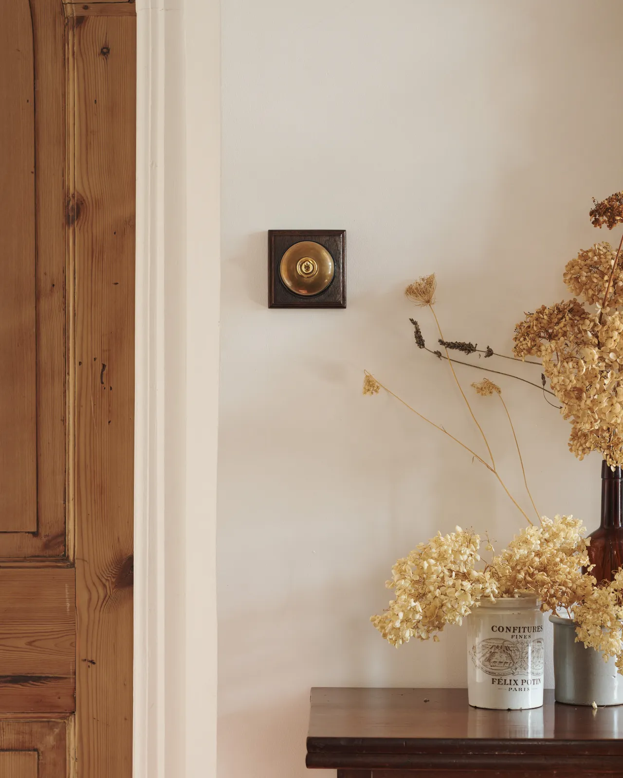 A deVOL Heirloom Brass Dolly Toggle Switch with a black ceramic base and richly stained Sapele pattress. Installed in a creamy coloured room styled with creamware pots and dried flowers.