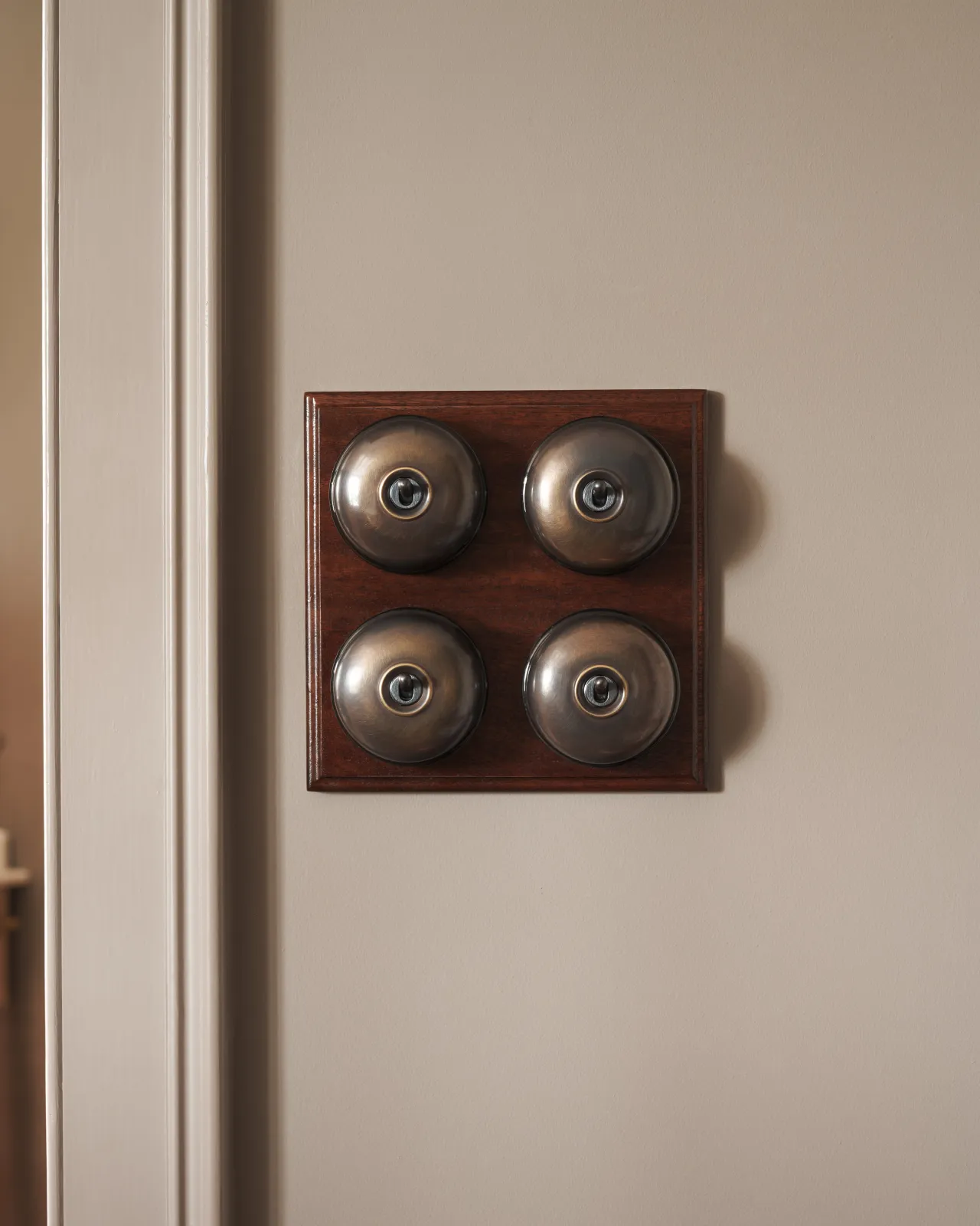 A bank of four Oxidised Brass Dolly Toggle Switches fitted to a richly stained Sapele pattress. Photographed in an atmospheric room, with warm-grey painted walls.