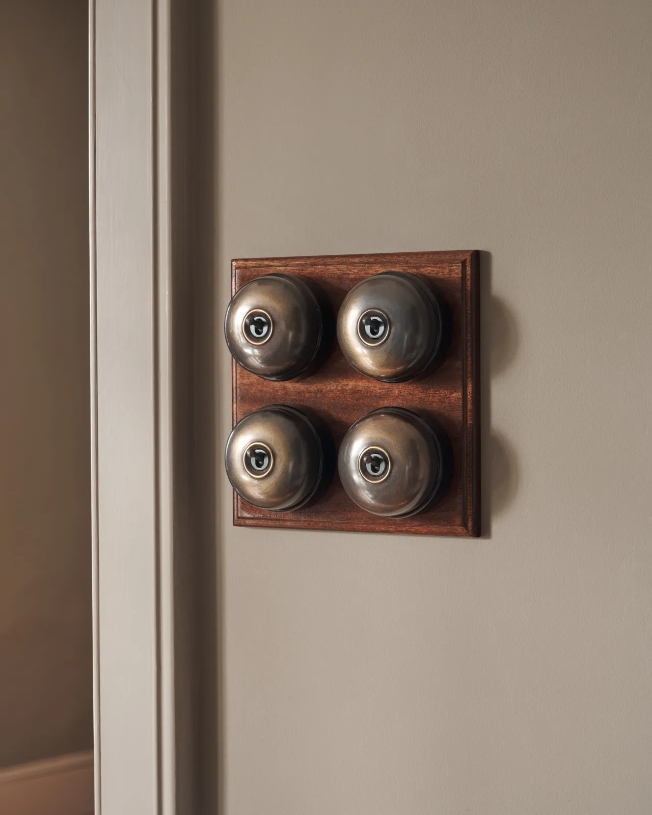 A bank of four Oxidised Brass Dolly Toggle Switches fitted to a richly stained Sapele pattress. Photographed in an atmospheric room, with warm-grey painted walls.
