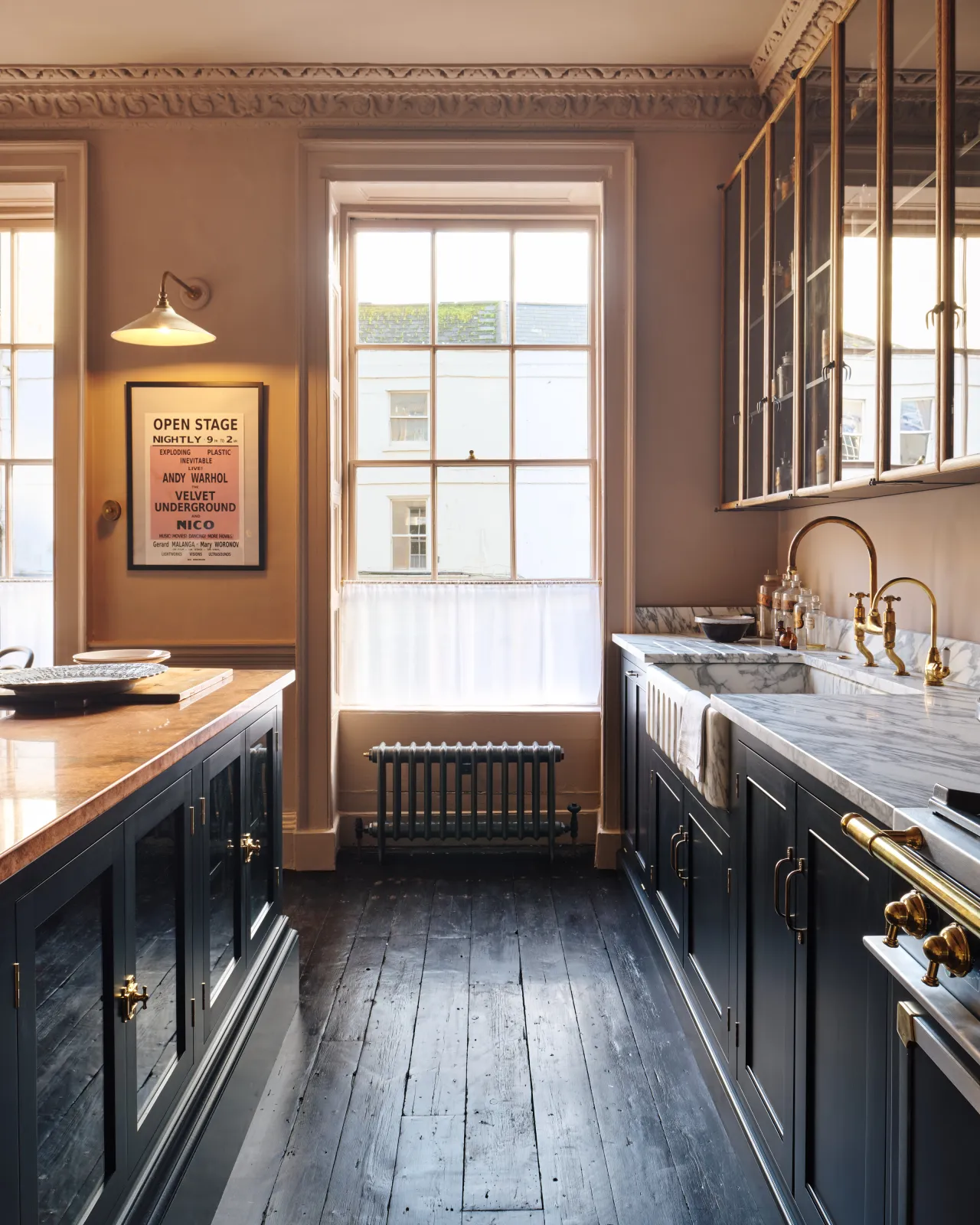 Looking between an island and Georgian-style kitchen sink cabinet with black painted cabinetry, an Arabescato marble sink, an aged brass tap, dark floorboards, pink walls and large original sash window.