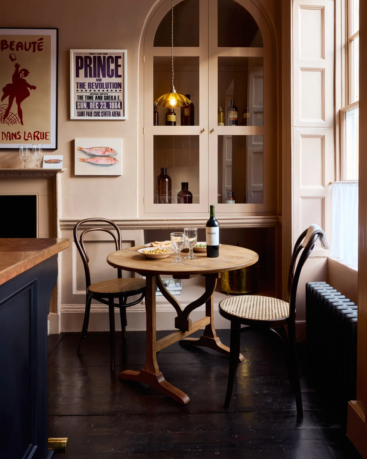 A straight-on view of a small round pine dining table in a dark Georgian-style room with pink walls, fitted glazed cupboard doors, black polished floorboards and vintage posters and styling.