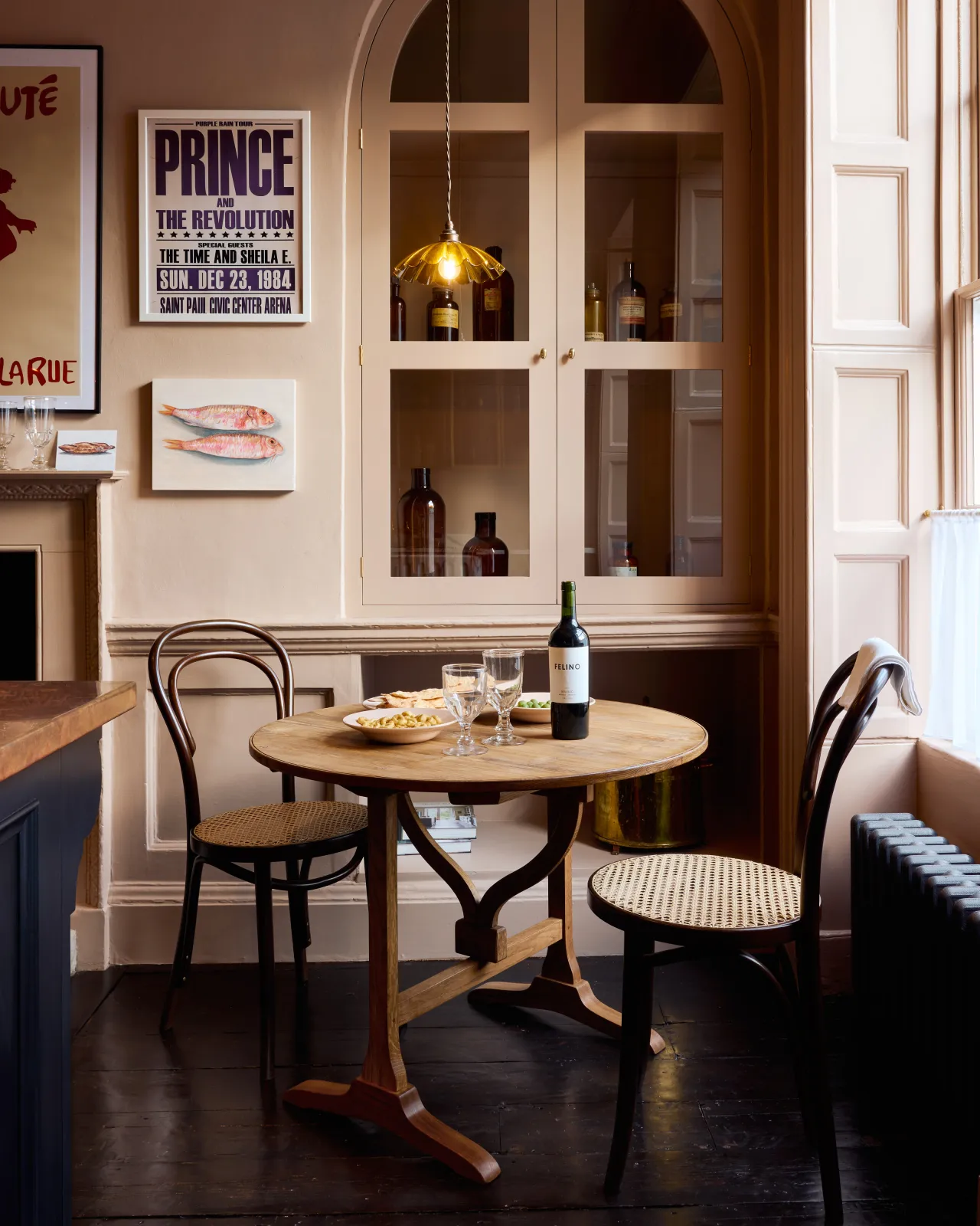 A straight-on view of a moody Georgian-style café-style corner with a small round pine dining table, pink walls and soft pink painted fitted glazed cupboard doors.