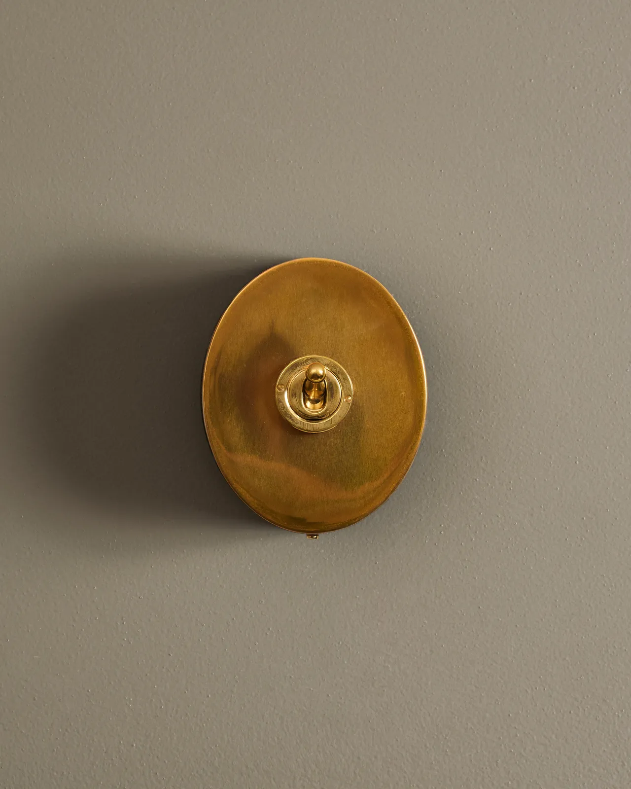 A straight-on view of a small 1 gang oval-shaped toggle switch in a warm burnished brass finish with a black base.