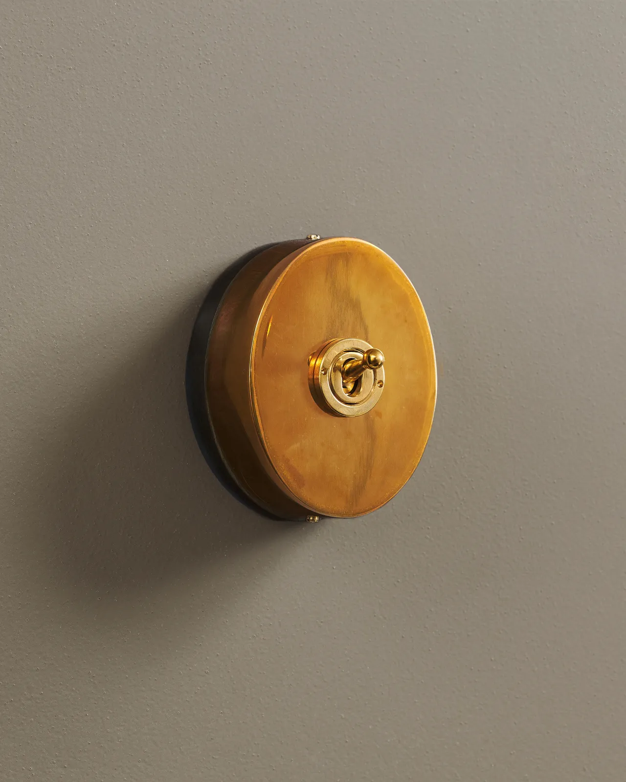 An oblique view of a small 1 gang oval-shaped toggle switch in a warm burnished brass finish with a black base.
