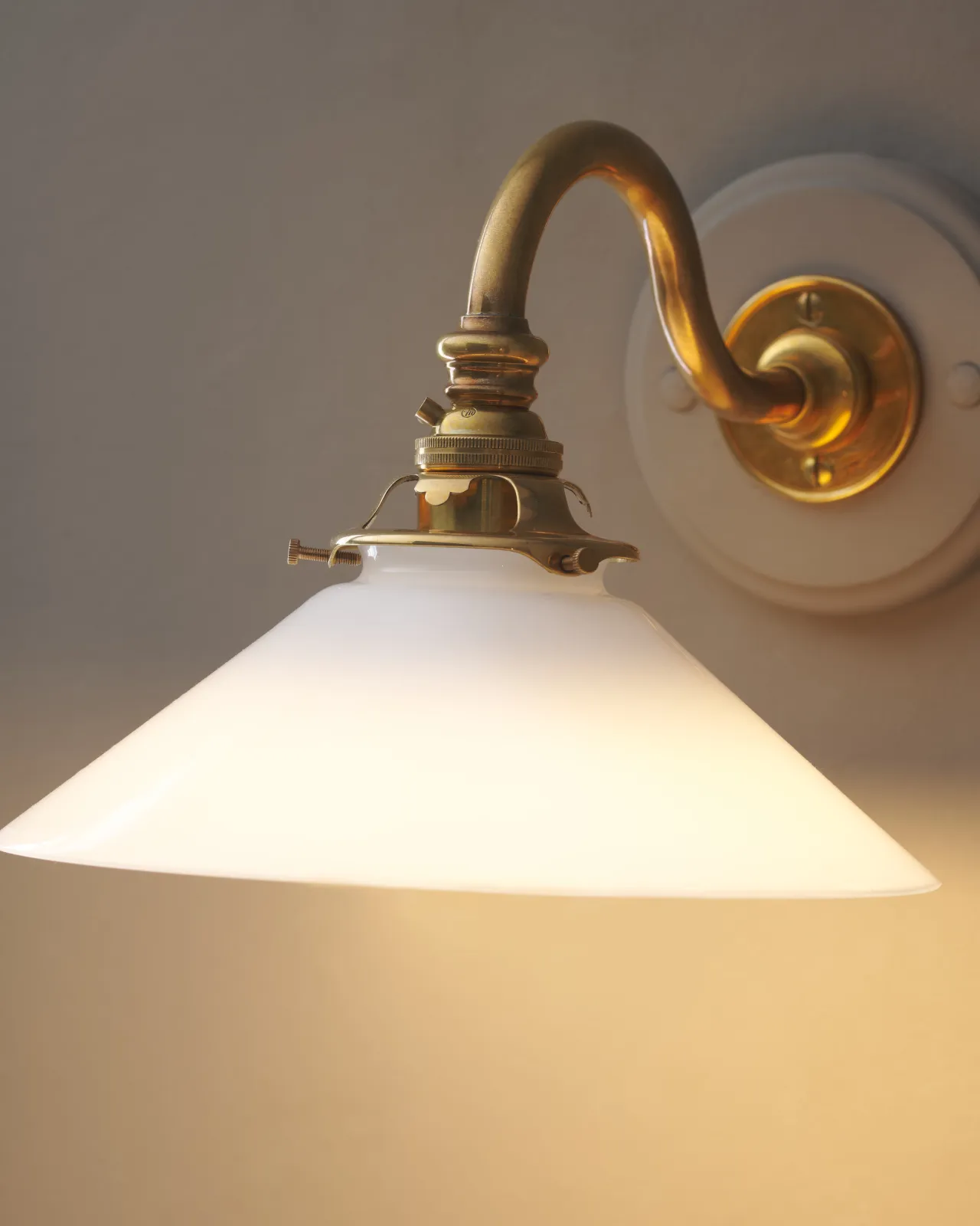 A close-up view of an elegant wall light with a curved brass arm, painted wooden pattress and small milk glass shade. A warm golden light glows through the glass shade.