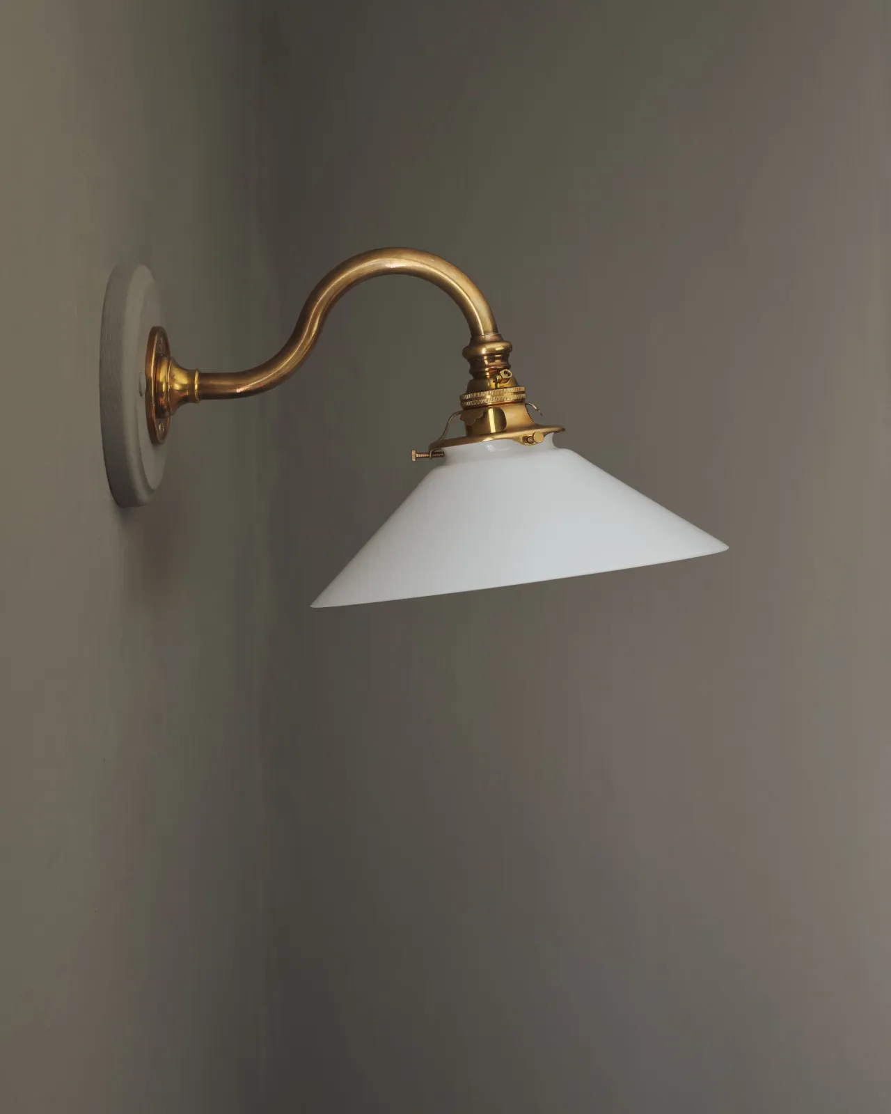 A side profile view of an elegant wall light with a curved brass arm, painted wooden pattress and small milk glass shade.