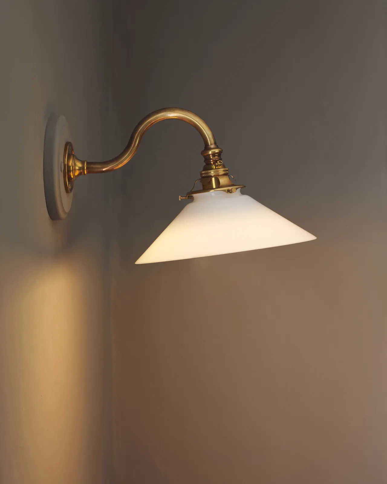 A side profile view of an elegant wall light with a curved brass arm, painted wooden pattress and small milk glass shade. A warm golden light glows through the glass shade.