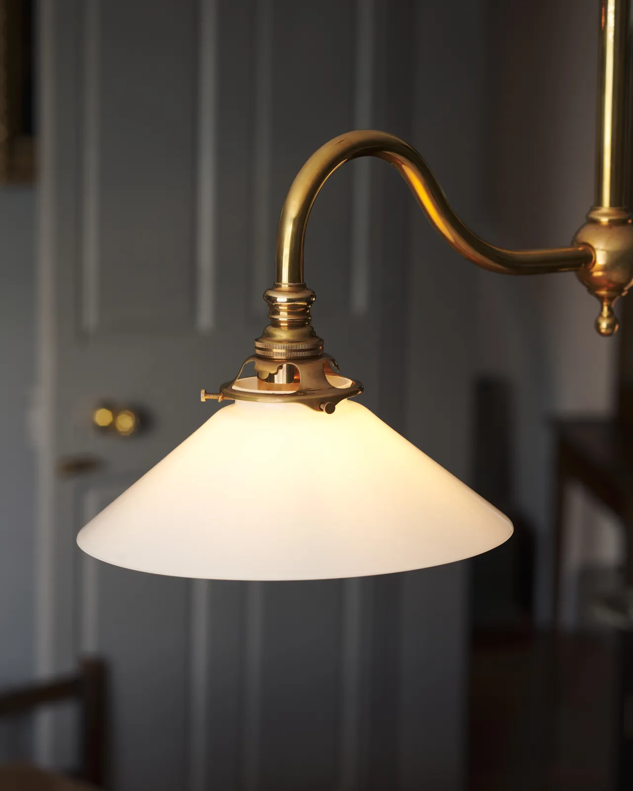 An illuminated small milk glass light shade connected to a curved brass arm via a delicate brass gallery with three thumbscrews.