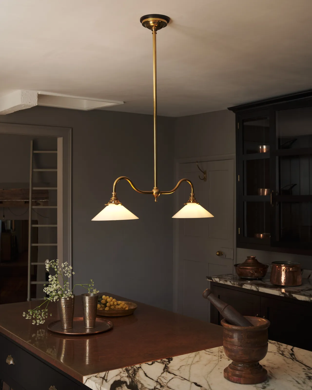 An elegant ceiling light with a rigid brass pole and two curved arms, each holding an illuminated small milk glass shade. The light gives off a warm glow above an electric and atmospheric dark kitchen.