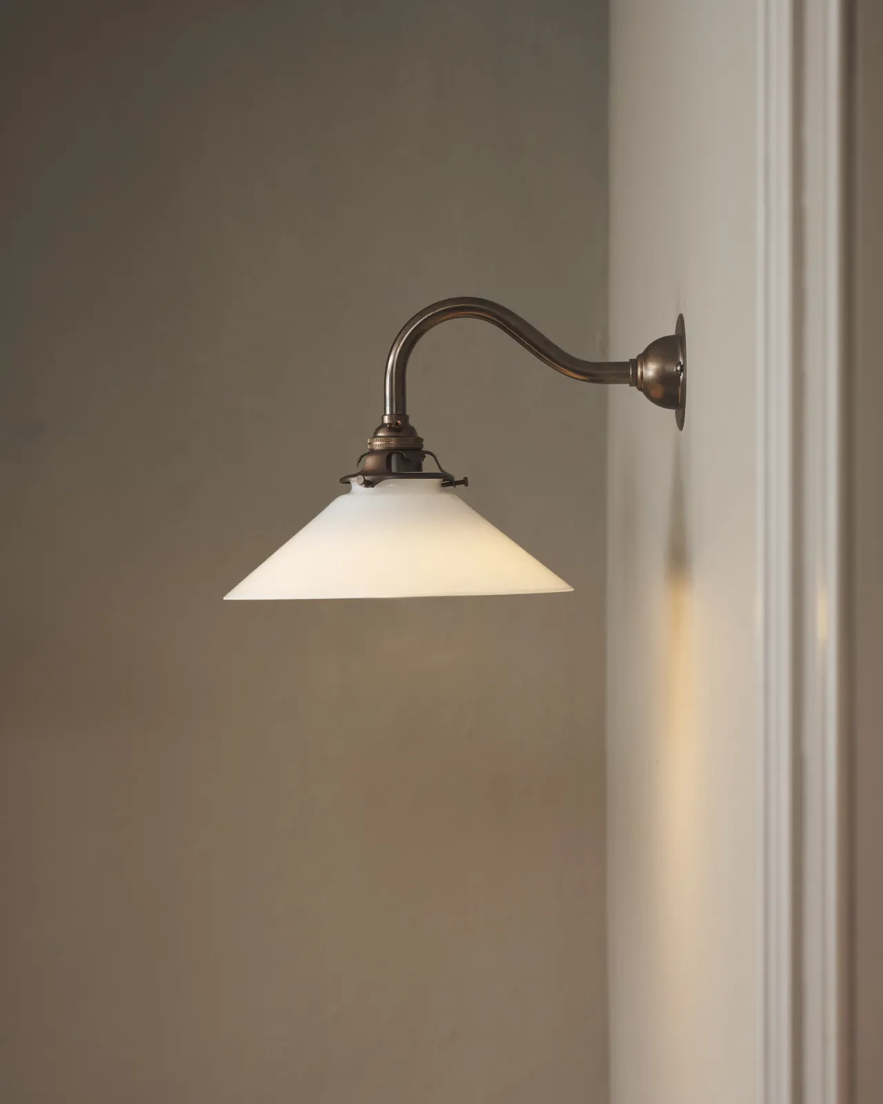 A simple swan neck wall light with an aged brass rose, arm and gallery holding a small illuminated milk glass shade.