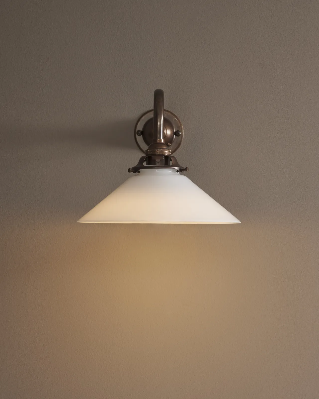 A close-up straight on view of a small milk glass wall light. The light is on and casting a warm glow across the wall.