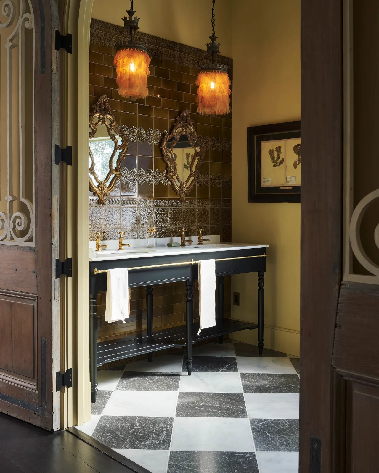 Looking through a doorway into a room with a black and white chequered marble floor and a classic black twin washstand with brass fittings and a wall of lace-patterned glossy sepia tiles.