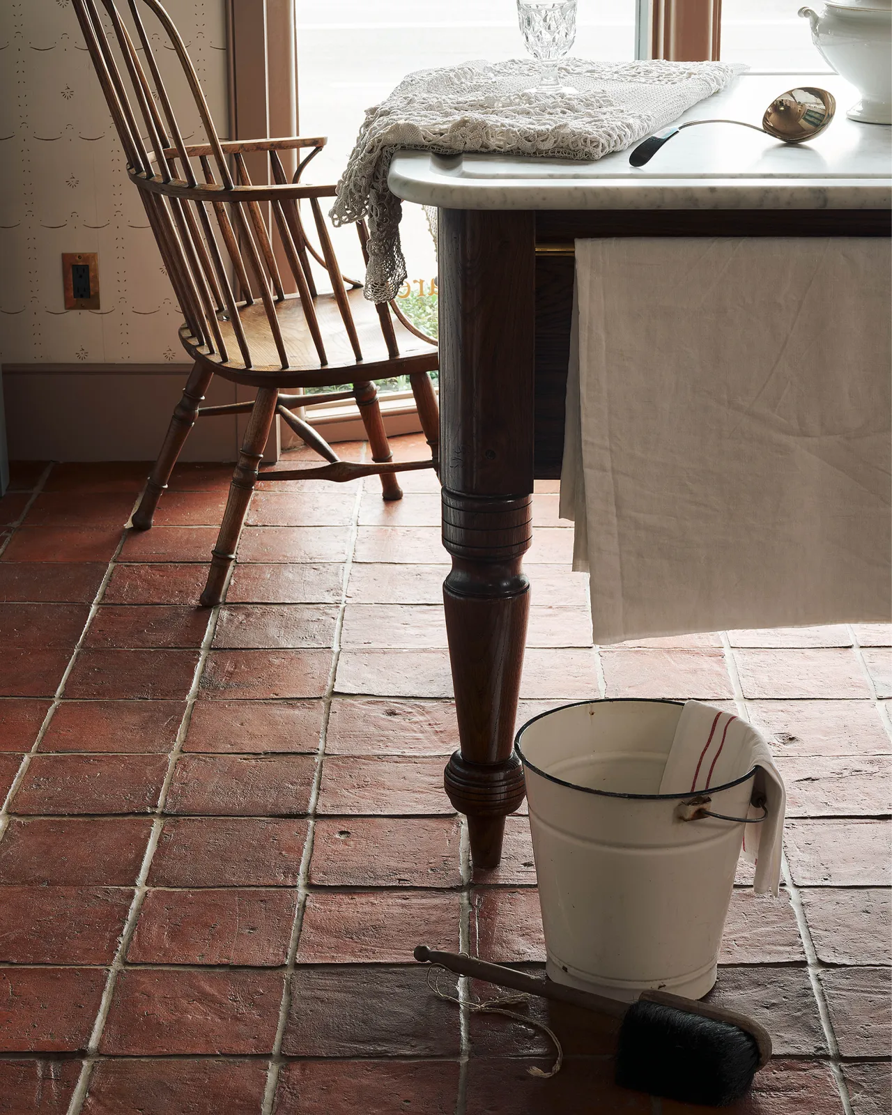 Light reflecting off a rustic terracotta tile floor, with an enamel bucket and vintage brush resting beside an elegant oak table leg.