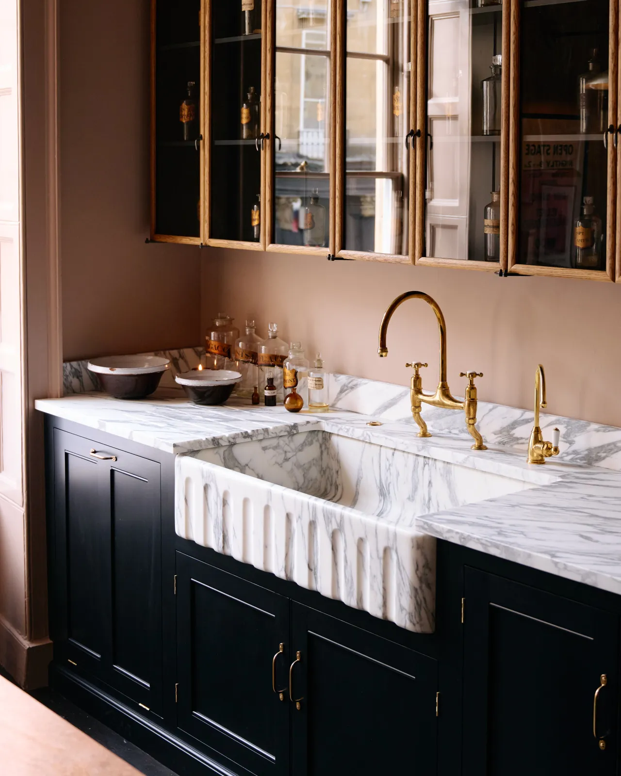 An oblique view of an extra large single bowl fluted Arabescato marble sink in a black painted Georgian-style kitchen with pink walls and mid-century-style glazed wall cupboards.