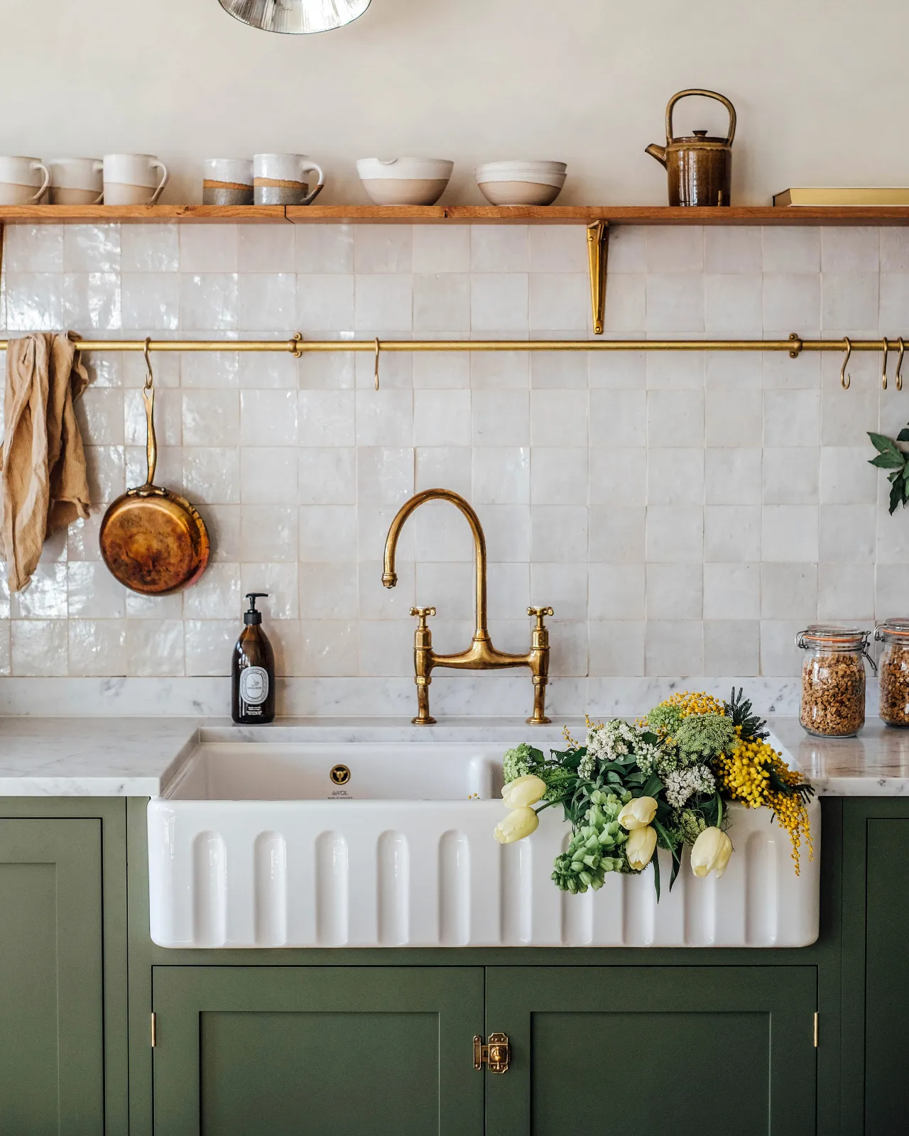 A straight-on view of an extra large double bowl fluted ceramic Belfast sink in a green painted Shaker-style kitchen with white metro wall tiles and an aged brass hanging rail.