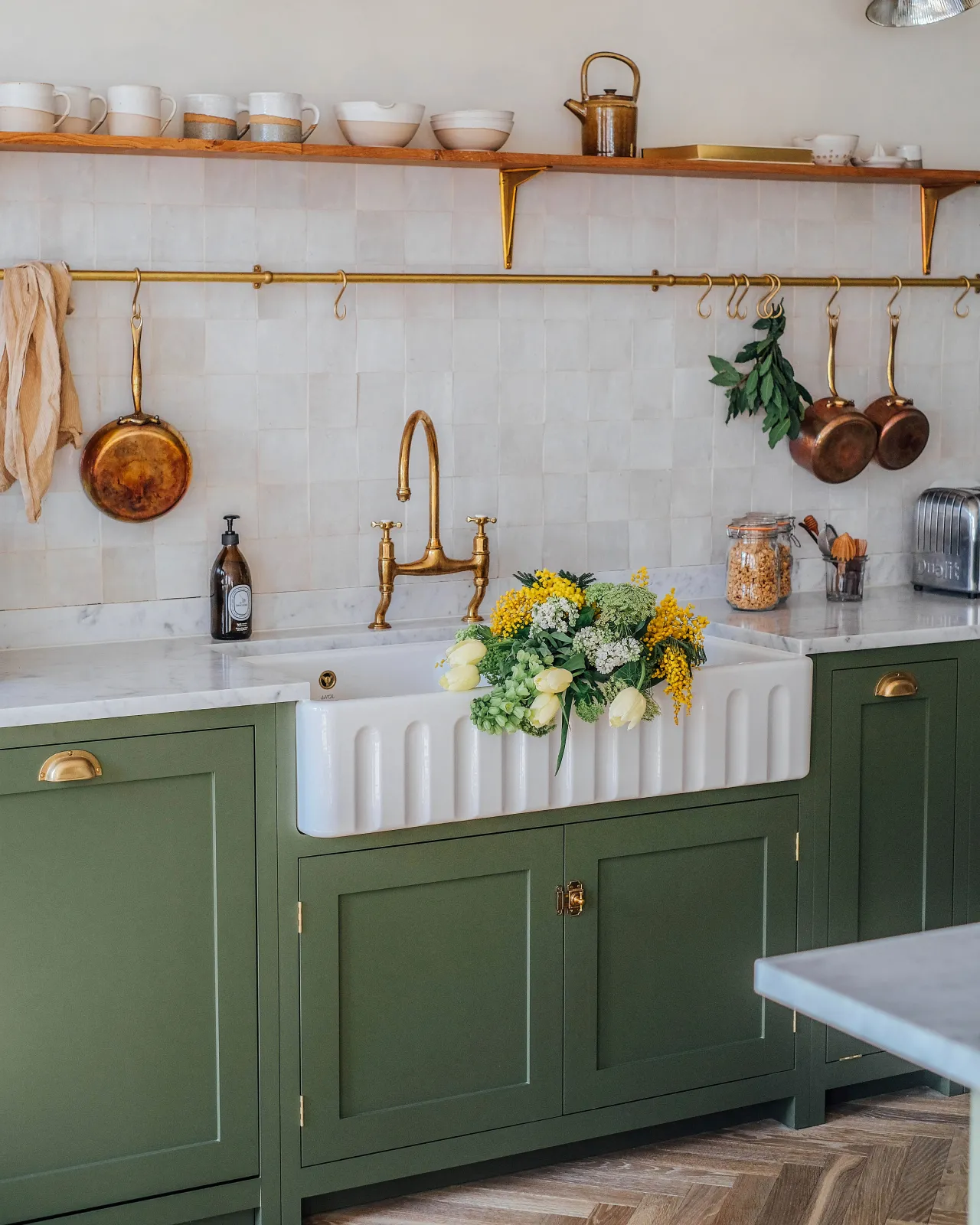 An oblique view of an extra large double bowl fluted ceramic Belfast sink in a green painted Shaker-style kitchen with white metro wall tiles and an aged brass hanging rail.