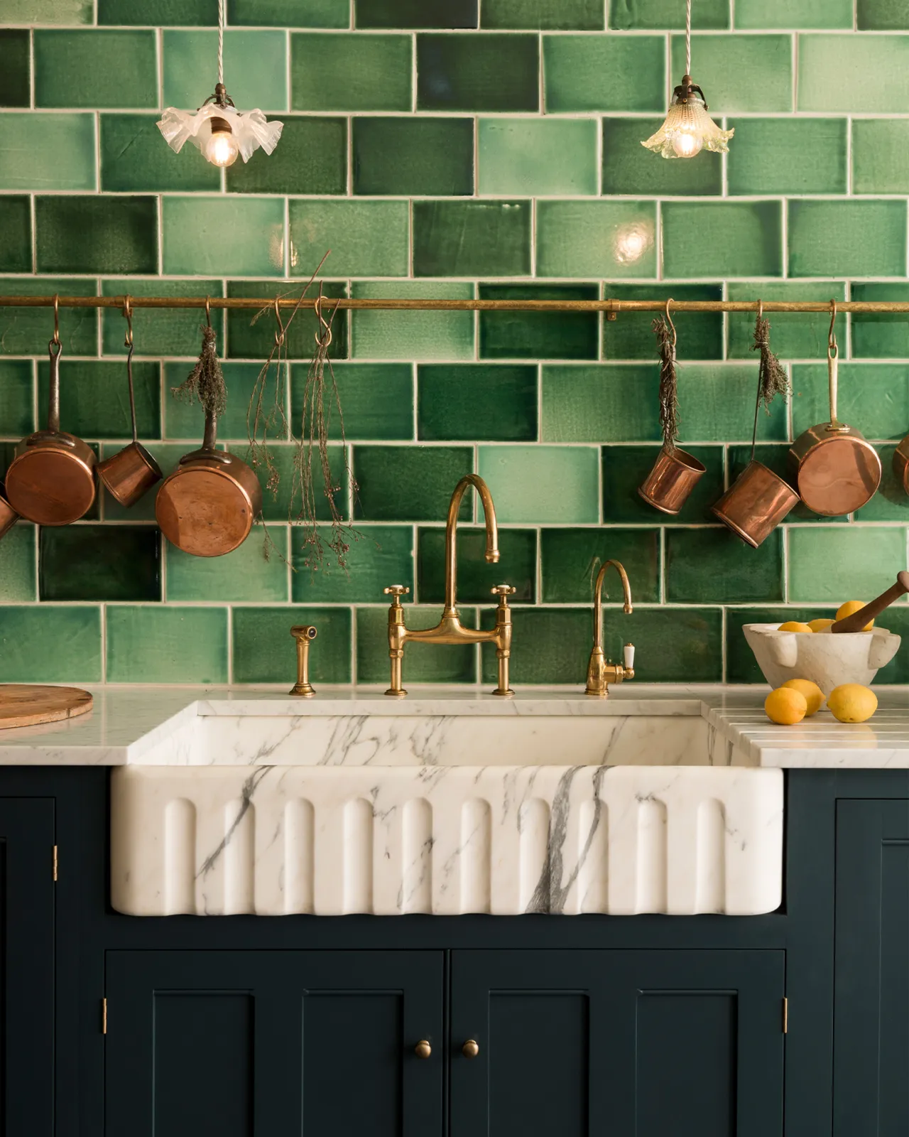A straight-on view of an extra large single bowl fluted Arabescato marble sink in a royal blue painted Georgian-style kitchen with glossy green wall tiles and a collection of vintage copper pans hanging from an aged brass hanging rail.