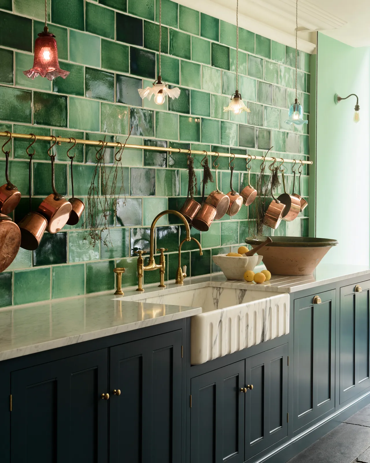 An oblique view of an extra large single bowl fluted Arabescato marble sink in a royal blue painted Georgian-style kitchen with glossy green wall tiles and a collection of vintage copper pans hanging from an aged brass hanging rail.