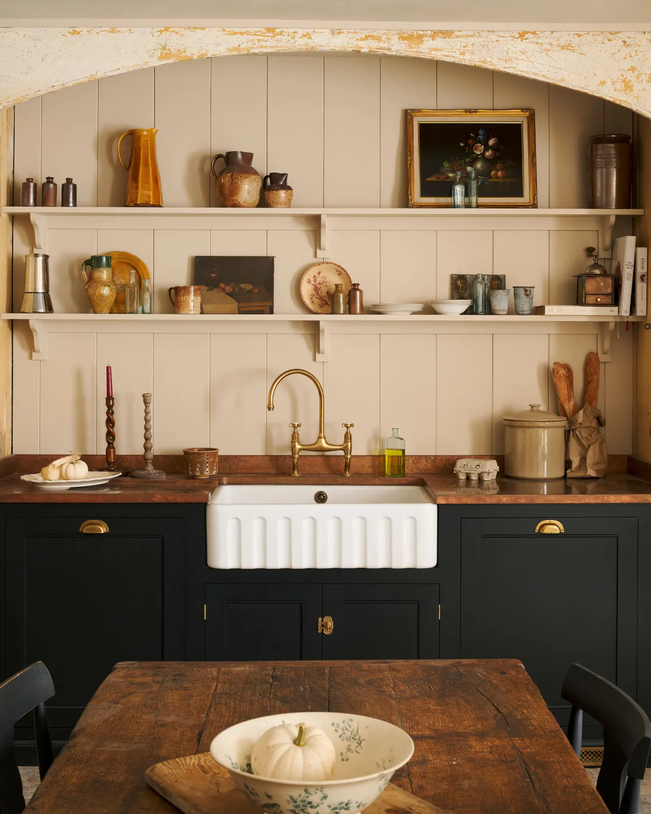 A straight-on view of a large fluted single bowl ceramic Belfast sink in a black painted Georgian-style kitchen with an aged brass mixer tap, an aged copper worktop and cream tongue and groove wall panelling with shelves full of vintage bric-a-brac.