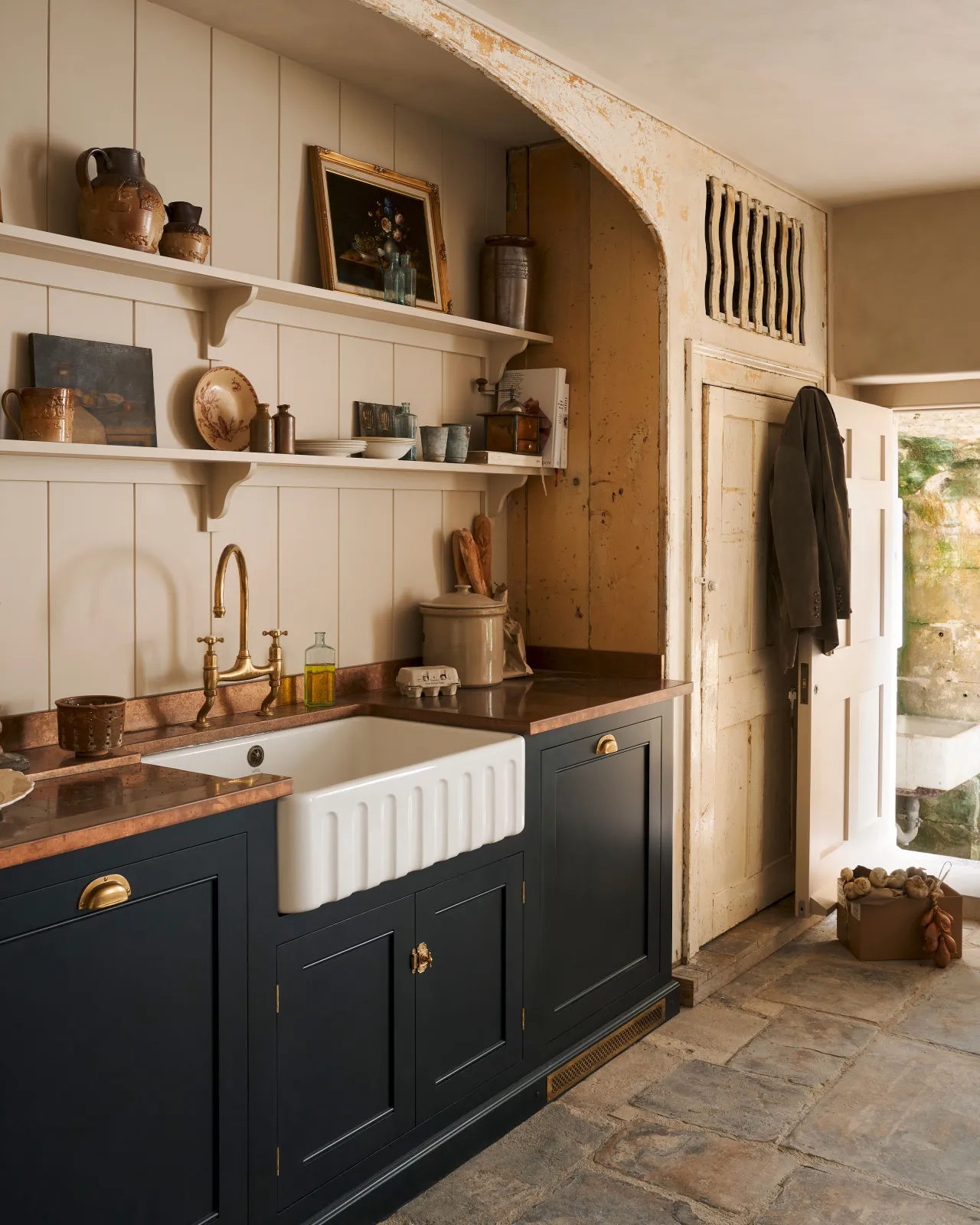 An oblique view of a large fluted single bowl ceramic Belfast sink in a black painted Georgian-style kitchen with an aged brass mixer tap, an aged copper worktop and cream tongue and groove wall panelling with shelves full of vintage bric-a-brac.