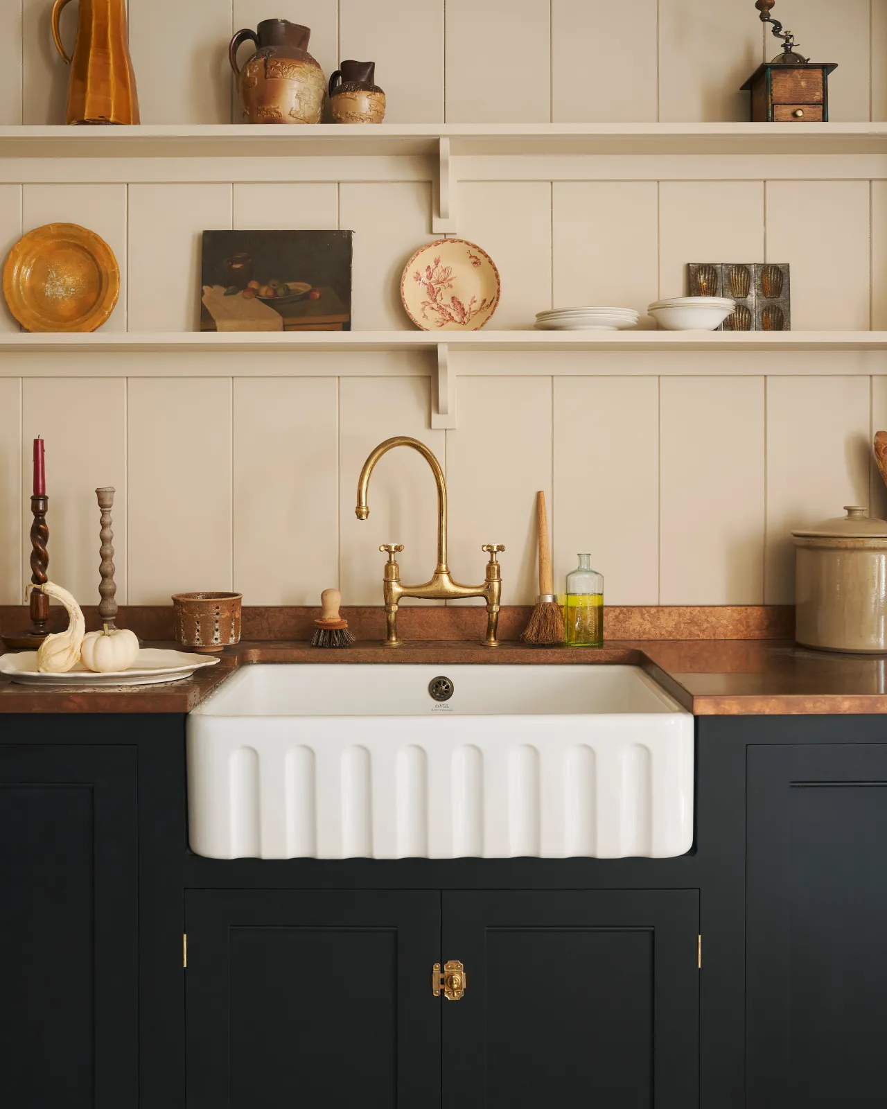 A close-up straight-on view of a large fluted single bowl ceramic Belfast sink in a black painted Georgian-style kitchen with an aged brass mixer tap, an aged copper worktop and cream tongue and groove wall panelling with shelves full of vintage bric-a-brac.