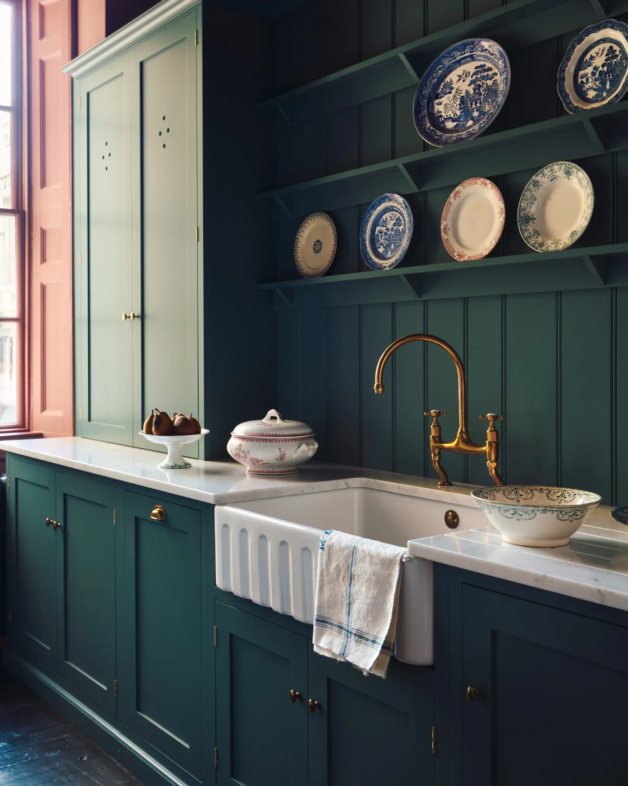 A close-up oblique view of a large fluted single bowl ceramic Belfast sink in a blue-green painted Shaker-style kitchen with wine coloured walls and tongue and groove wall panelling with shelves decorated with vintage crockery.