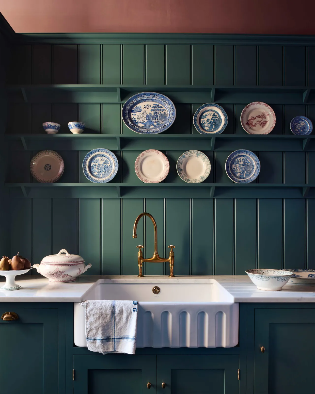 A close-up straight-on view of a large fluted single bowl ceramic Belfast sink in a blue-green painted Shaker-style kitchen with wine coloured walls and tongue and groove wall panelling with shelves decorated with vintage crockery.
