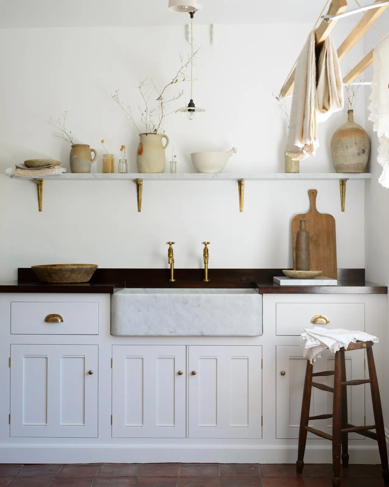 A straight-on view of a large single bowl Carrara marble sink in a white painted Georgian-style kitchen sink cabinet with a dark hardwood worktop, white walls and terracotta flooring.
