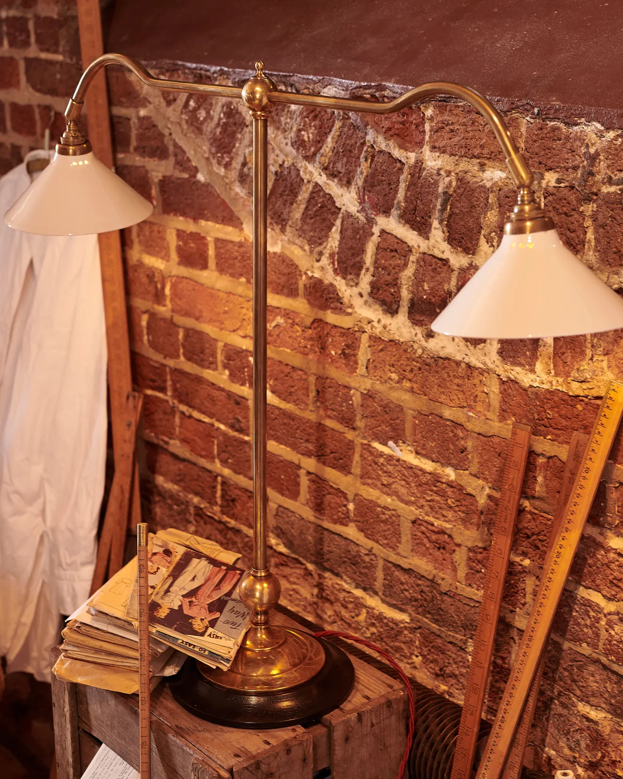 An oblique view of a Victorian-inspired desk lamp with a solid brass foot, stand, two curved arms and two small cream glazed ceramic light shades on a wooden box in front of a red brick wall.