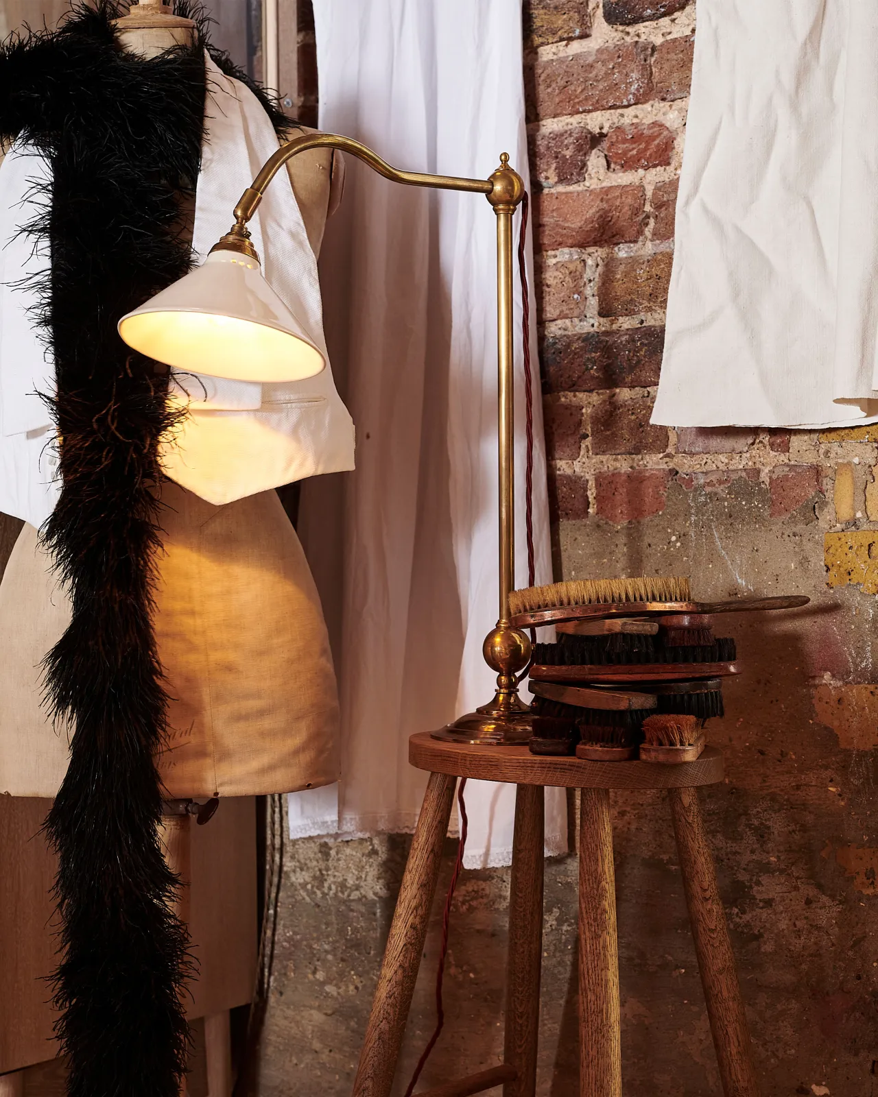 A Victorian-inspired desk lamp with a solid brass foot, stand, curved arm and a small cream glazed ceramic light shade, sitting on a simple oak bar stool against a red brick wall.