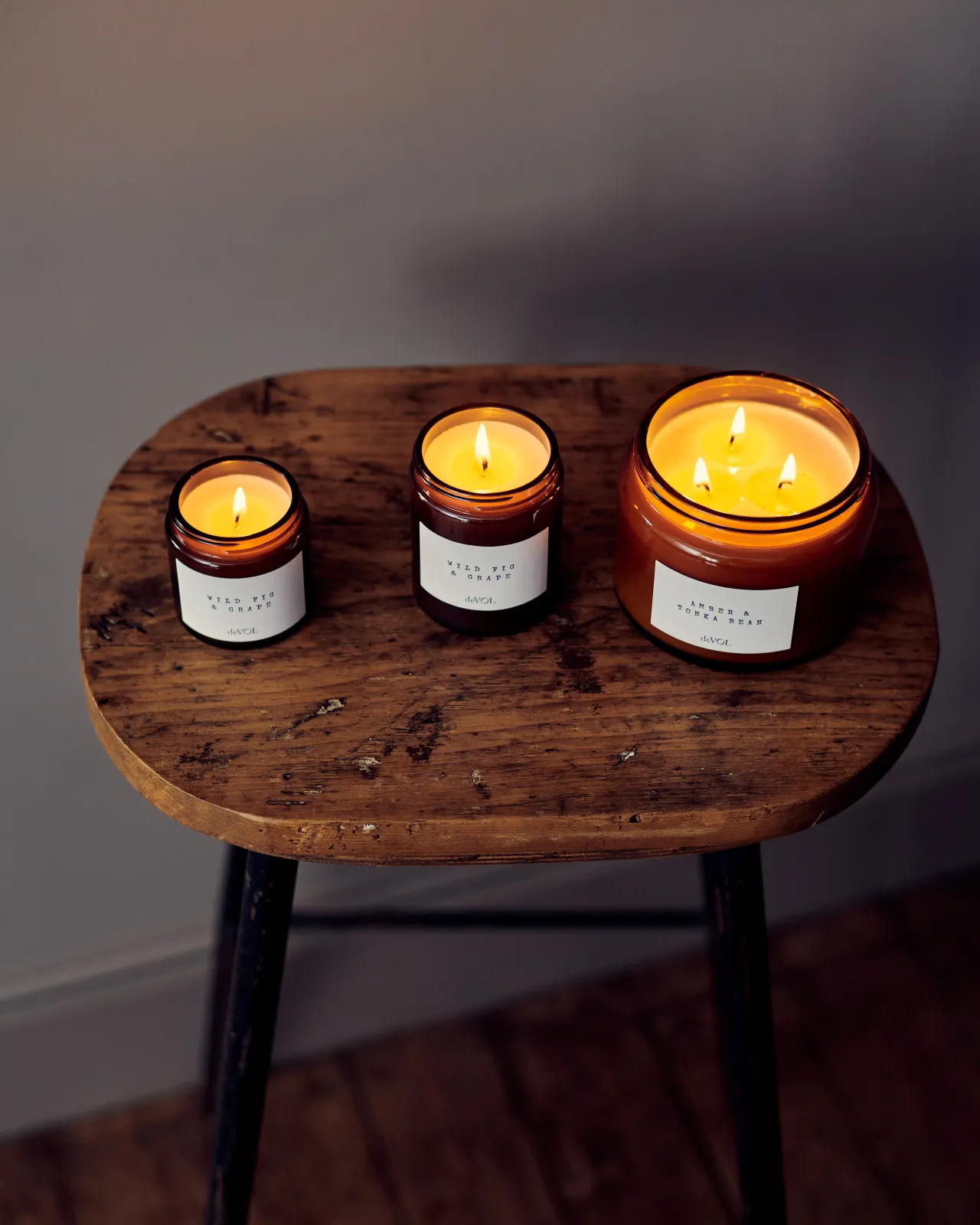 An overhead view of a collection of three lit scented candles in brown glass jars on a wooden stool top; small, medium and large.