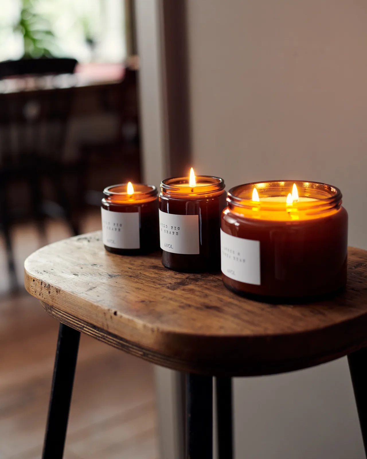 An oblique view of a collection of three lit scented candles in brown glass jars on a wooden stool top; small, medium and large.