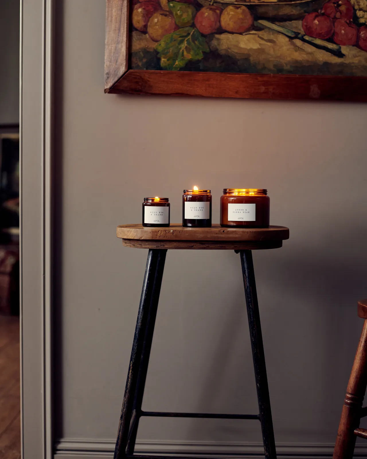 A straight-on view of a collection of three lit scented candles in brown glass jars on a wooden stool top; small, medium and large.