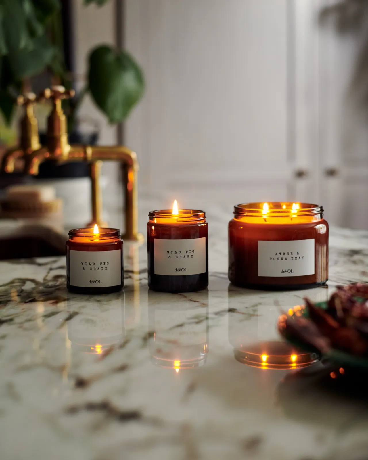 A collection of three lit scented candles in brown glass jars on a polished quartz worktop; small, medium and large.