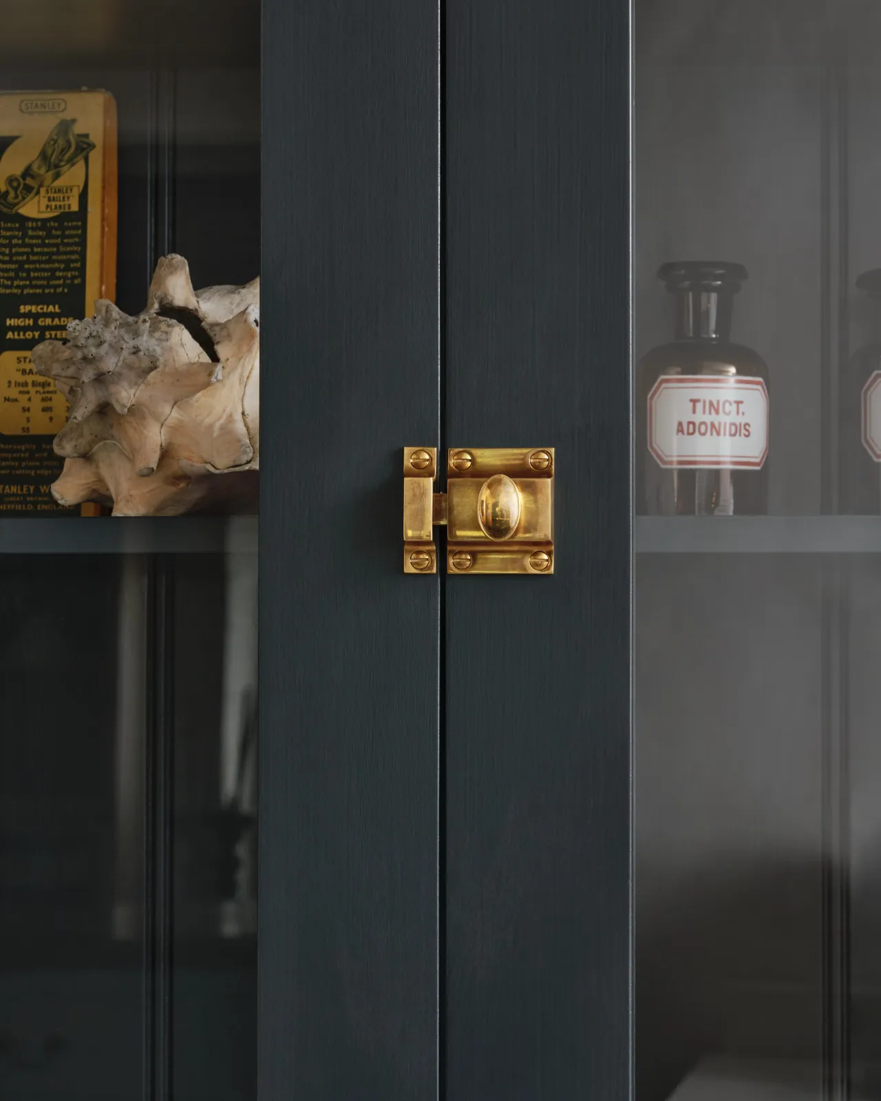 A straight-on view of a classic-style square cupboard catch with an ornate oval-shaped knob in a warm burnished brass finish fitted to dark blue painted glazed cupboard doors.