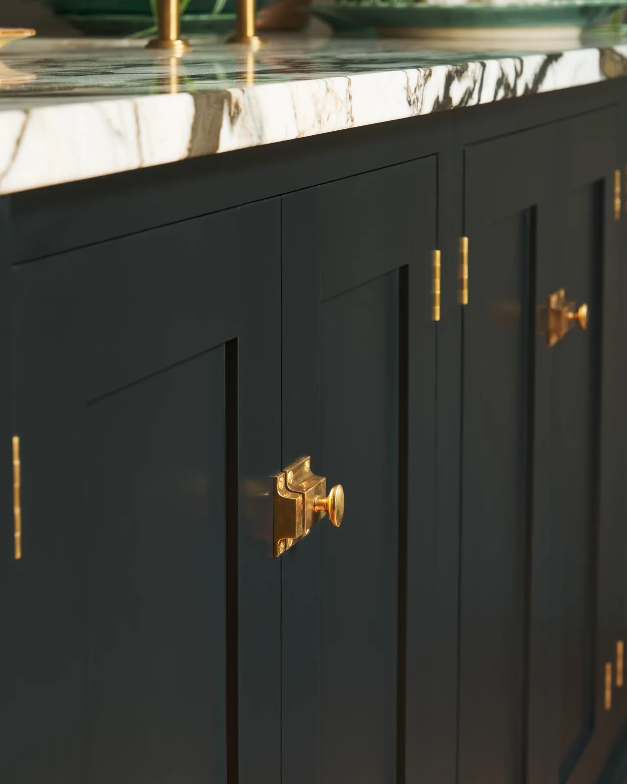 An oblique view of classic-style square cupboard catches in a warm burnished brass finish fitted to black painted kitchen cupboard doors.