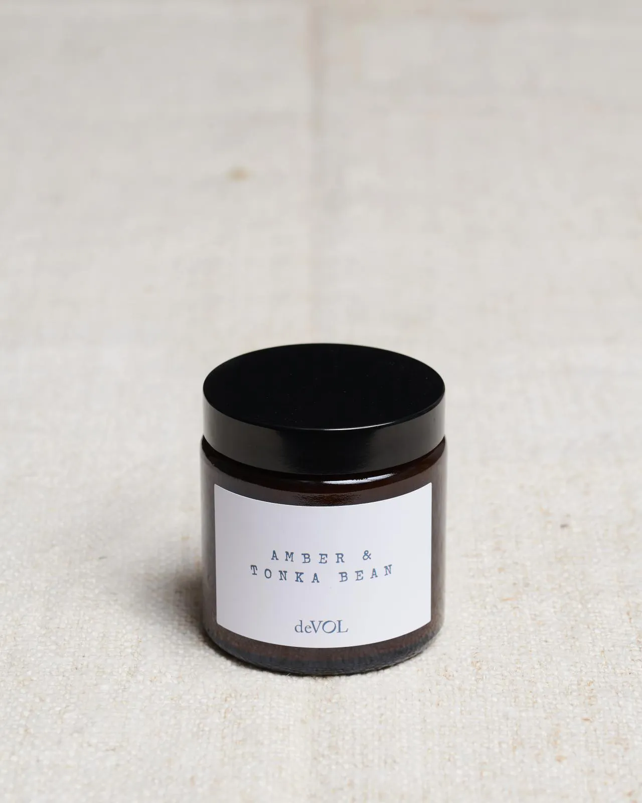 A small scented candle in a brown glass jar with a black lid, the label reads 'Amber & Tonka Bean, deVOL'.