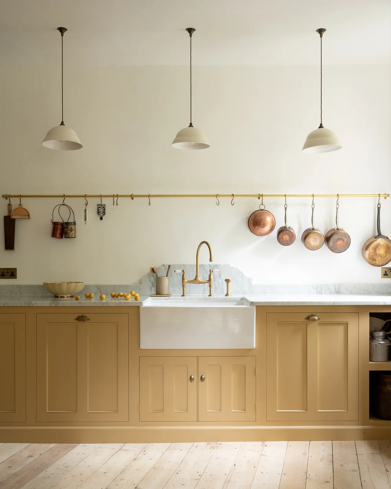A straight-on view of a yellow painted Georgian-style sink cupboard an aged brass hanging rail fixed to the wall above. Several vintage copper pots and utensils hang via brass s hooks along the rail.