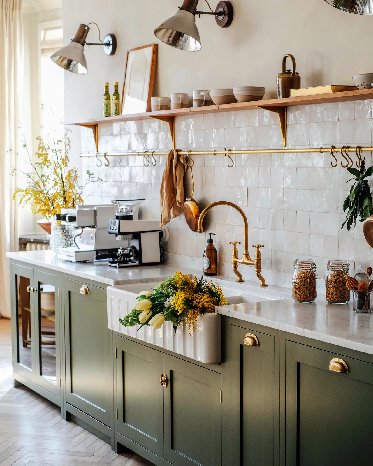 An oblique view of a Shaker-style sink cupboard with a fluted Belfast sink, aged brass mixer tap and an aged brass hanging rail fixed to white metro tiles. Several vintage copper pots and linen tea towels hang via brass s hooks along the rail.