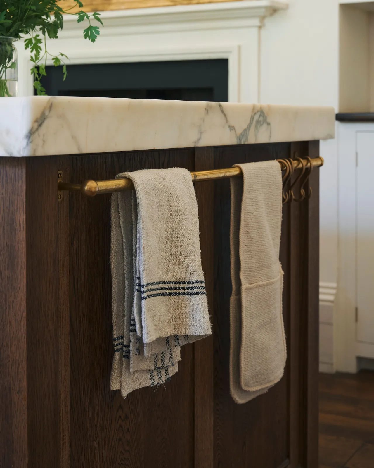 An oblique view of a short aged brass hanging rail fixed to the end of a kitchen island. Two vintage linen tea towels are draped over the rail.