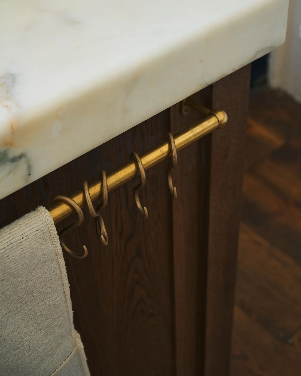A close-up oblique view of a short aged brass hanging rail fixed to the end of a kitchen island. A collection of brass s hooks hang along the rail.