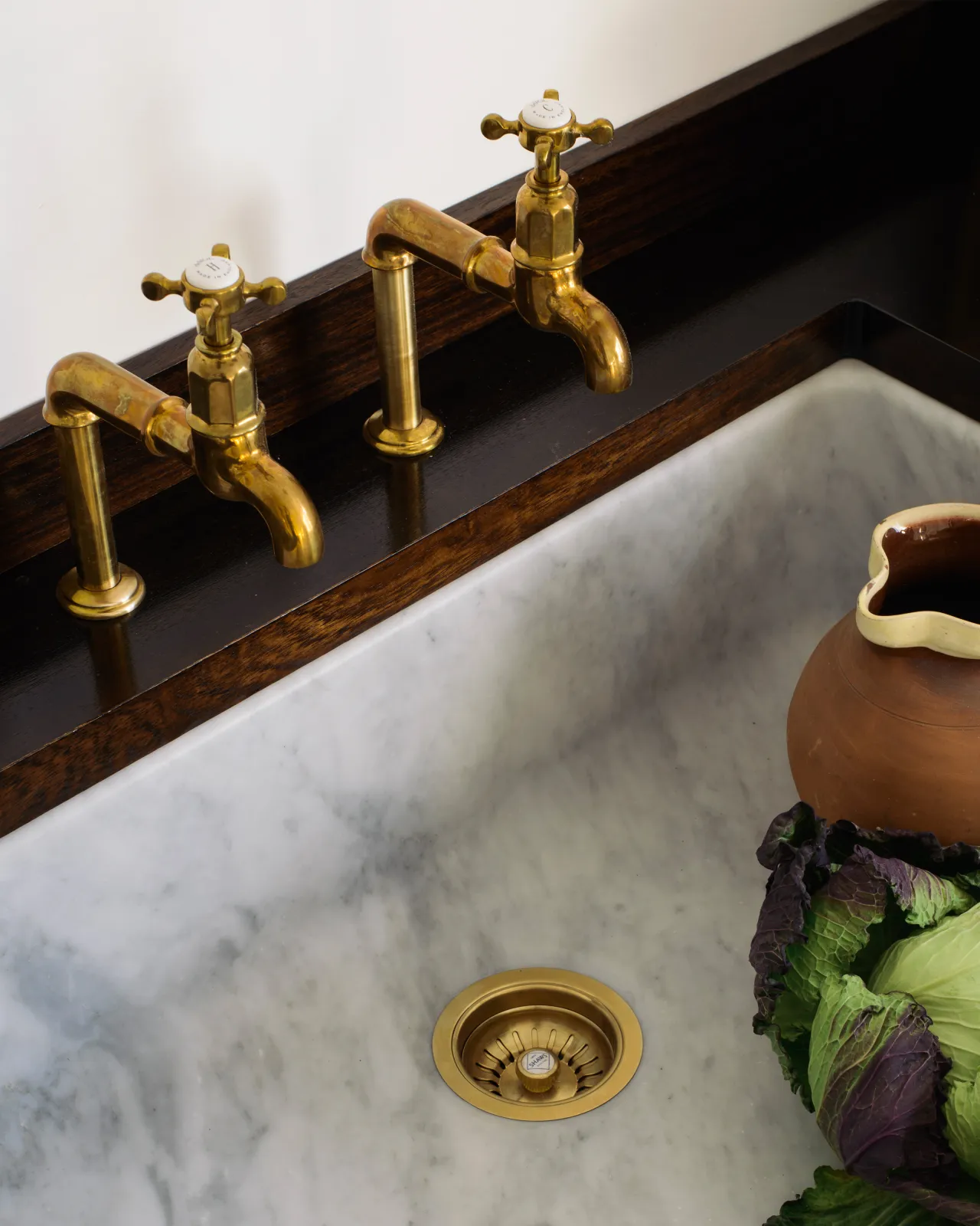 A pair of traditional-style aged brass bibcock taps with crosshead handles seen from overhead. They are fitted to a dark hardwood worktop over an elegant Carrara marble sink.