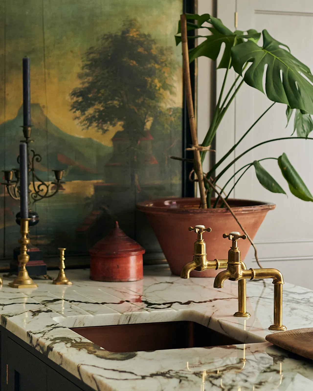 A profile view of a pair of traditional-style aged brass bibcock taps with crosshead handles, seen in a bohemian-style kitchen with a dramatic quartz worktop, a monstera plant and a large antique oil painting.
