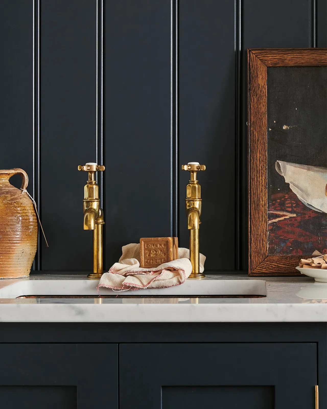 A straight-on view of a pair of traditional-style aged brass bibcock taps with crosshead handles in a classic utility kitchen with a Carrara marble worktop and deep blue painted cupboards and tongue and groove panelling.
