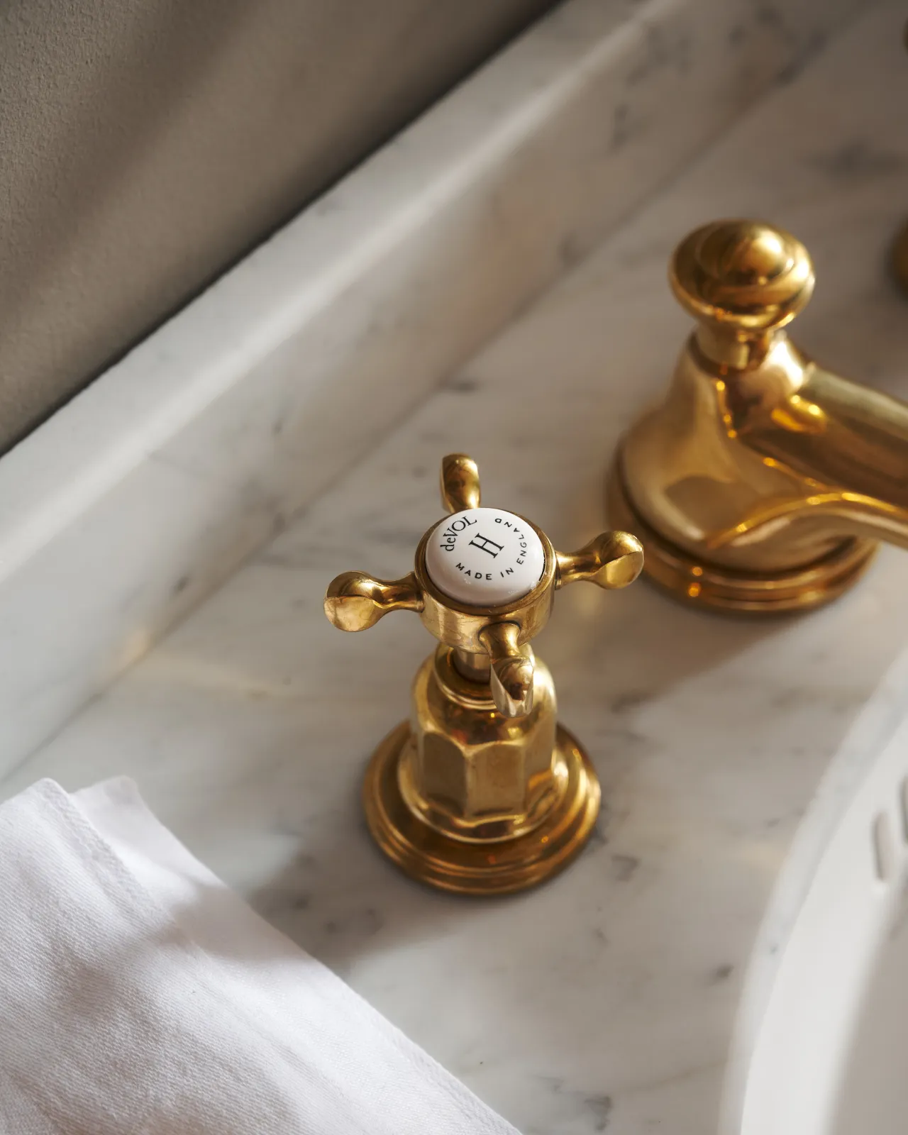 A close-up oblique view of the crosshead handle of an aged brass mixer tap for a bathroom basin sink. The letter 'H' is in the centre of the handle top, surrounded by the words 'deVOL, Made in England'.