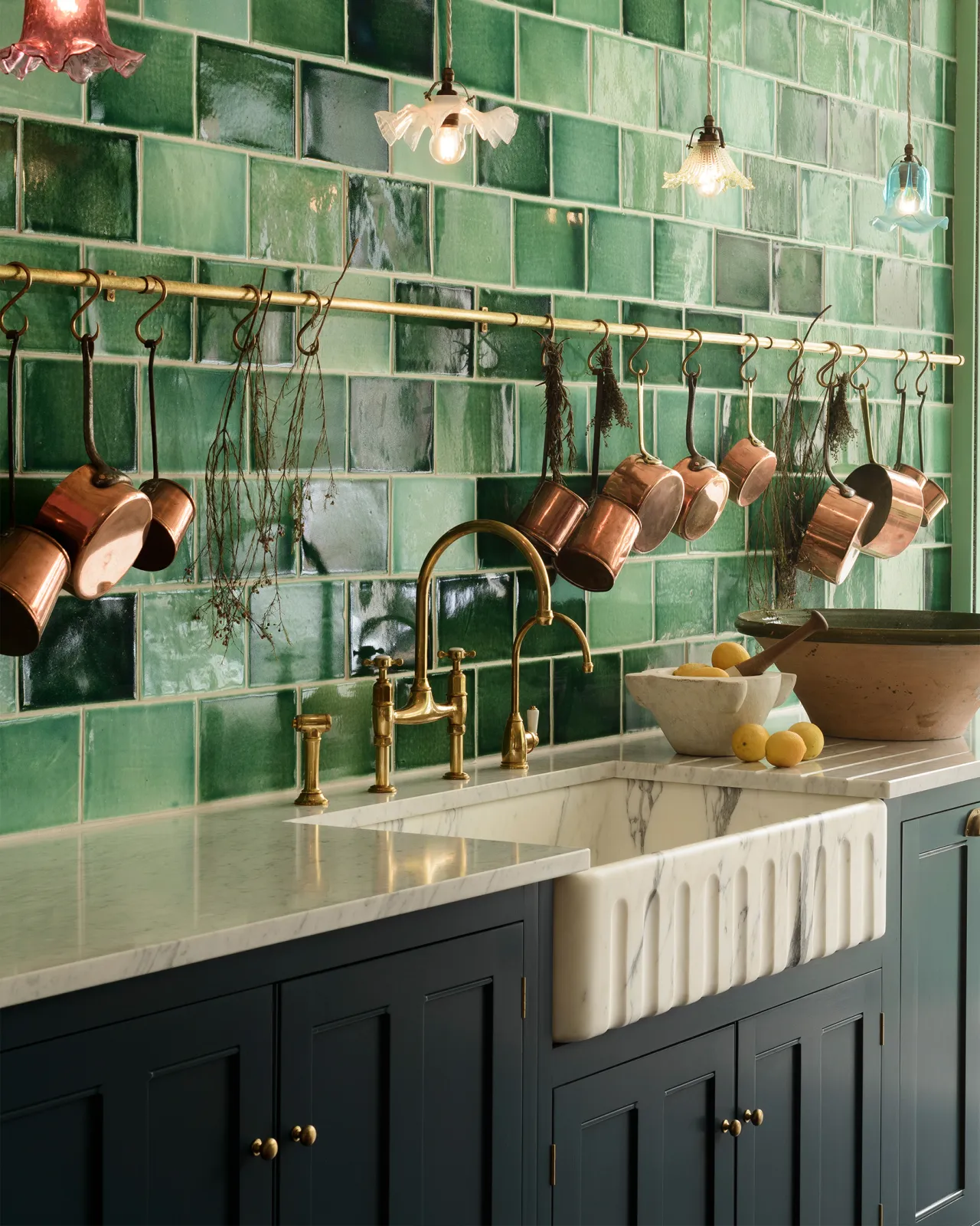An oblique view of a Georgian-style kitchen sink with an Arabescato marble sink and a wall of glossy green wall tiles. A long aged brass hanging rail is fixed above the sink with a large collection of vintage copper pans hanging from brass s hooks.